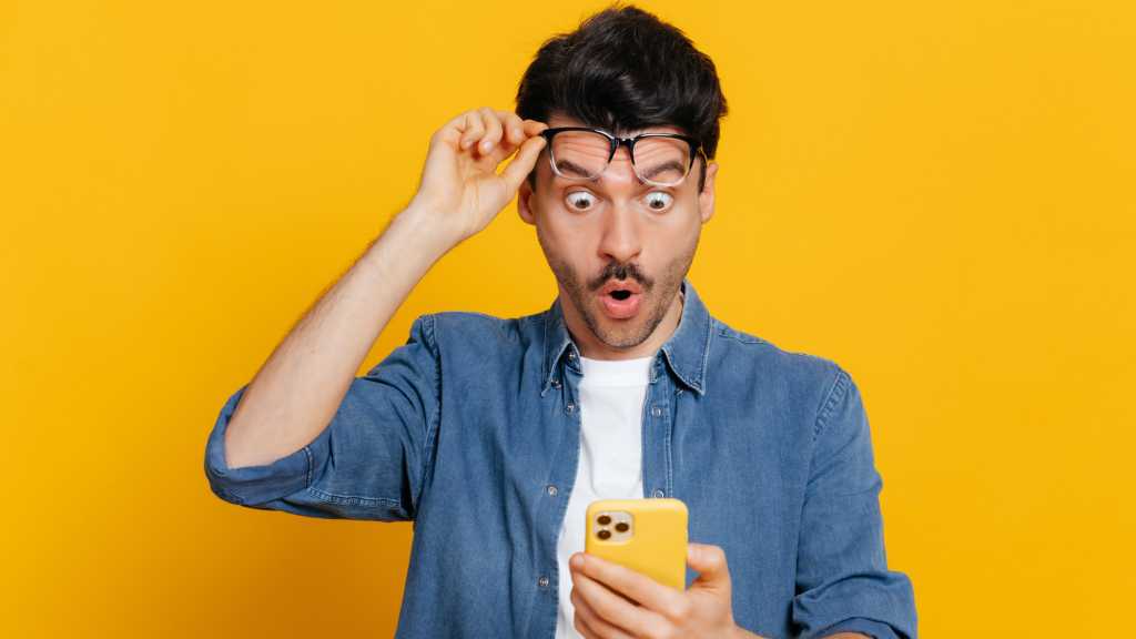 Mann schiebt Brille hoch und starrt überrascht auf sein gelbes Handy