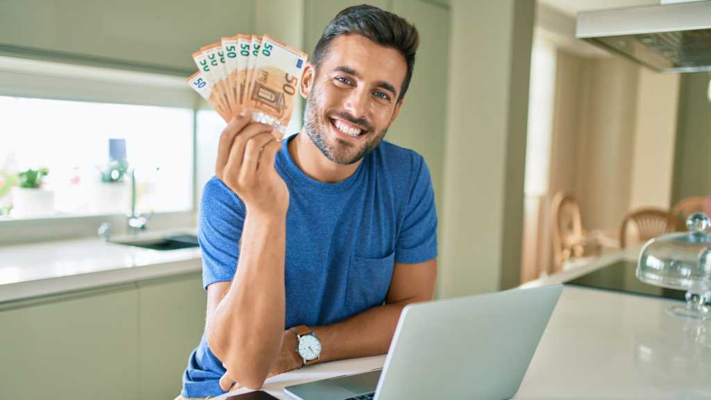 Geld sparen im Internet: Bärtiger junger Mann hält in der rechten Hand mehrere 50-Euro-Geldscheine. Vor ihm steht auf dem Tisch ein Notebook.